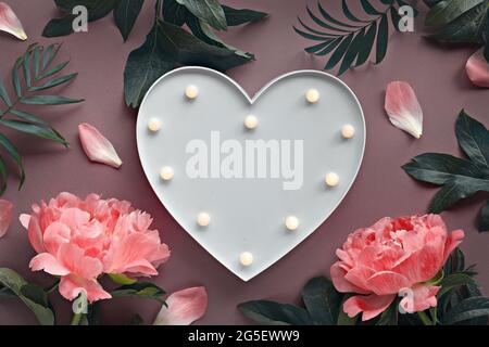 Creative flat lay with light coral peony and exotic palm leaves around heart shaped light board on rosy brown or dark beige background. Stock Photo