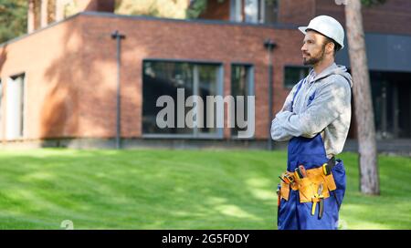 Young bearded male engineer in hard hat looking away, posing outdoors while working on cottage construction Stock Photo