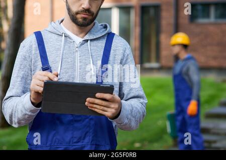 Cropped shot of young male builder holding digital tablet pc with coworker working in the background at cottage construction site Stock Photo