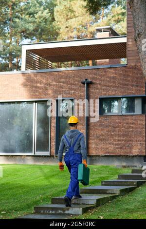 Full length shot of young male builder in blue overalls carrying toolbox at cottage construction site Stock Photo