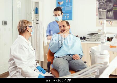 Sick patient complaining about toothache while talking with dentist before intervetion. Senior doctor speaking to man sitting on stomatological chair while nurse stomatology hospital office Stock Photo