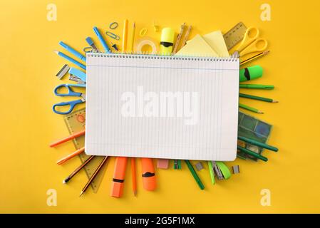 Educational notebook with blue lines open on pile of school supplies on yellow background. Top view. Stock Photo