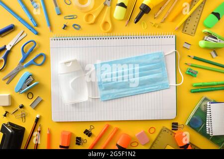 New school year with protection against covid-19 with mask and gel on notebook and tools around on yellow background. Top view. Stock Photo