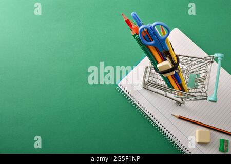 Purchase of school tools with cart full of supplies on notebook on green background. Top view. Stock Photo