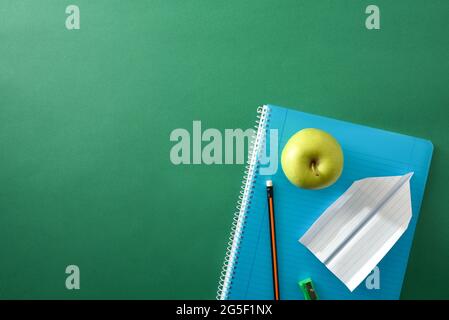 Blue spiral notebook with pencil, sharpener, green apple and paper plane on top on green background with space for text. Top view. Stock Photo