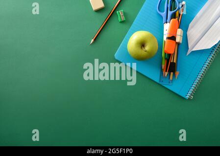 Blue spiral notebook with pencil, sharpener, green apple and paper plane on top on green background with space for text. Top view. Stock Photo