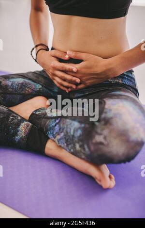 Cropped shot of woman sitting on yoga mat with her legs crossed and hands over her belly. Female doing yoga at fitness class. Stock Photo
