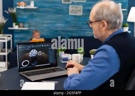 Senior engineer working on deadline sitting at desk. Experienced engineer working from home office on mechanical project using laptop computer technology and wife is reading a book on sofa. Stock Photo