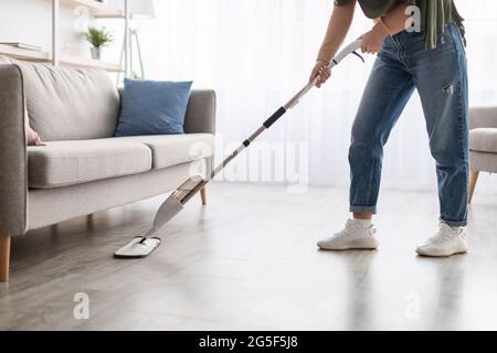 Chores Concept. Closeup cropped low section of islamic woman in headscarf washing hardwood laminate flooring under sofa using water spray mop pad and Stock Photo