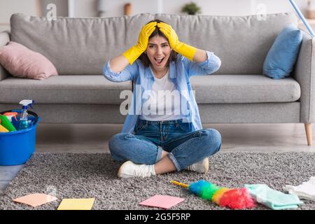 Angry woman screaming tired of cleanup, wearing rubber gloves and grabbing head, sitting on floor surrounded by cleaning equipment, taking break. Exha Stock Photo