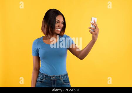 Happy Black Young Woman Making Selfie On Smartphone Posing Standing On Yellow Studio Background. Social Media, People And Gadgets Concept. Modern Life Stock Photo