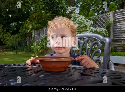 A sticky white Caucasian small boy age nearly 3 with curly blond hair sits outdoors with a grubby brown mouth eating chocolate cake Stock Photo