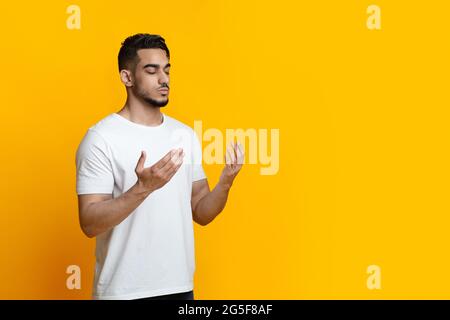 Religious young arab muslim man with closed eyes praying, holding palms face up, whispering pray, yellow studio background, panorama with copy space. Stock Photo