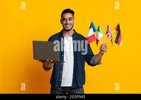 Cheerful arab guy student holding laptop and bunch of international flags, attending online class, yellow studio background, panorama. Foreign languag Stock Photo