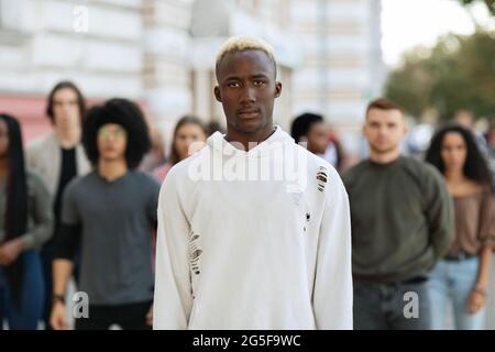 Serious african american young man standing over multiracial group of strikers on the street, black guy leading millennial activists, protesting again Stock Photo