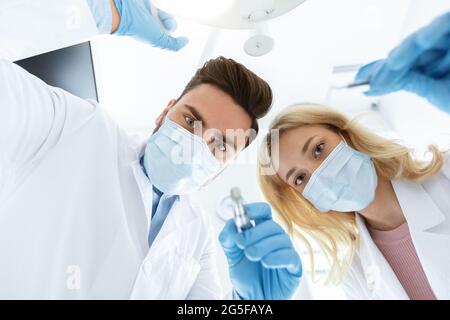 Two dentists young caucasian man and woman in face masks and rubber gloves doing treatment, turning on lamp, shot from dental chair. Modern stomatolog Stock Photo