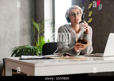 White-haired focused woman in headphones drinking coffee and eating cake in office Stock Photo