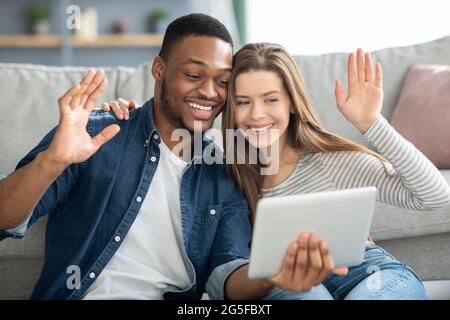 Web Chat. Happy Interracial Couple Holding Digital Tablet And Waving Hands At Webcamera, Cheerful Multicultural Lovers Making Video Call With Friends Stock Photo