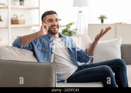 Distance Communication Concept. Portrait of excited smiling young israeli man talking with friends or family on mobile phone sitting on the couch, ges Stock Photo