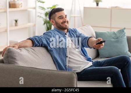 Leisure And Entertainment Concept. Smiling jewish guy watching TV show or film, holding remote control. Young man in kippa enjoying free time sitting Stock Photo