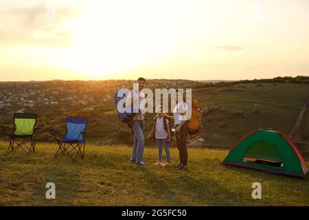 Happy family on summer camping trip at mountains. Adorble girl and parents standing at campground during sunset Stock Photo