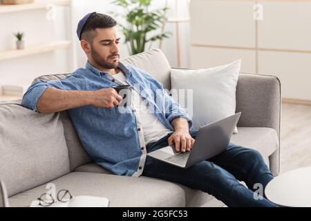 Portrait of focused handsome young jewish guy wearing yarmulke using personal computer holding mug and drinking coffee or tea. Man sitting on sofa and Stock Photo