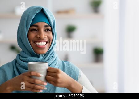 Portrait Of Positive Smiling Black Islamic Woman In Hijab Drinking Coffee At Home, Cheerful Young African Muslim Lady In Headscarf Holding Cup With Ho Stock Photo