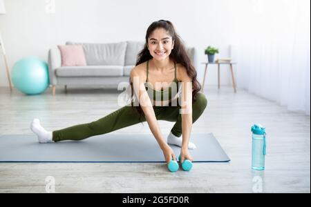 Young Indian woman doing stretching exercises with dumbbells on mat in ...