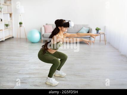 Home workout with virtual reality. Young Indian woman in VR headset doing squats at home. Athletic millennial Asian lady using modern technologies for Stock Photo