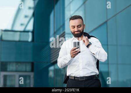 Business communication. Stylish mature businessman texting on cellphone walking outdoor against glass office center, empty space. Man using cellphone Stock Photo