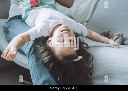 Dad and little girl 2-4 play on the couch. Kid lies on father's lap with her head down and laughs Stock Photo