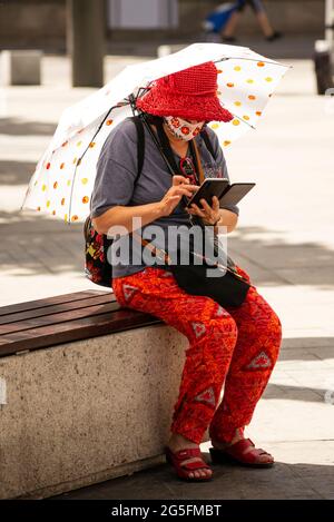 Lone senior woman wearing face mask, colourful cloths and umbrella using smartphone while sitting on a bench in urban environment Stock Photo