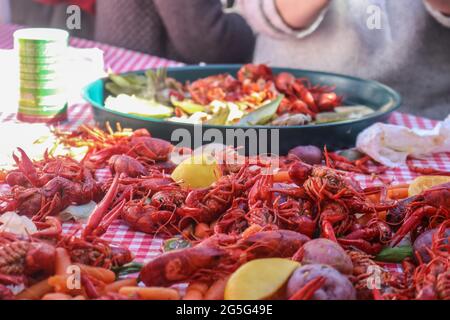 Boiled crawfish and vegetables piled on red checked tablecloth with eating tray and arm of person eating bokeh behind - shallow focus Stock Photo