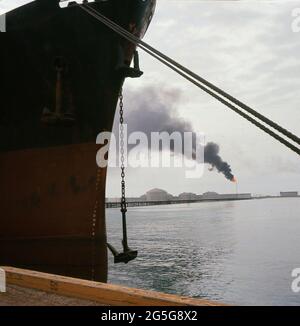 1960s, a ship moored in the oil port of the industrial area known as Ras Tanura, Saudi Arabia. Developed by the Arabian American Oil Company (Aramco) after the discovery of nearby oil in the 1930s, it grew to become a major Persian Gulf terminal of pipelines and artificial docks to accomodate large oil tankers. A petroleum refinery and storage tanks are also located there. Seen in the distance is a tall stack with a flame burning on top. This stack, called a flare, is a prominent feature of every oil refinery as it is an essential part of the plant's safety system. Stock Photo