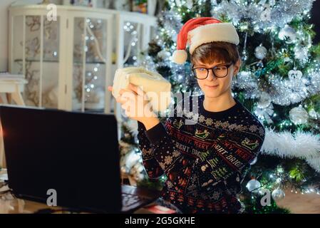 Young man with a red Santa hat holding gift box during online family greeting at home at Christmas time with Garlands lights background. Virtual house Stock Photo