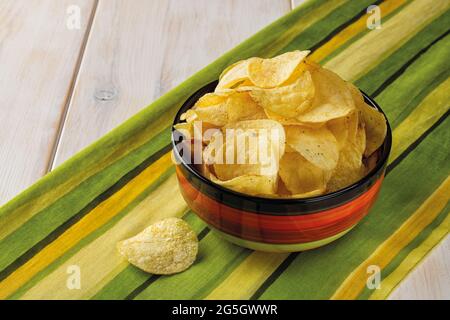 Crispy potato chips with dill in a ceramic bowl over striped yellow green table napkin. Popular snack of potatoes deep-fried thin slices. Fast food. Stock Photo