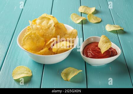 Delicious potato chips with dill and thick tomato ketchup in white ceramic bowls over wooden table. Crispy snack of potatoes deep-fried thin slices. Stock Photo