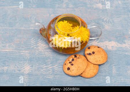Glass transparent teapot with healthy herbal tea from yellow dandelions and oatmeal cookies with raisins on a blue wooden background. Healthy lifestyl Stock Photo