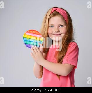 Happy kid girl in pink t-shirt and headband demonstrates, shows her new trendy sensory toy, rainbow color round - pop it Stock Photo