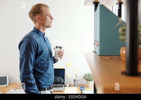 Young pensive businessman standing with cup of take out coffee and looking through office window Stock Photo