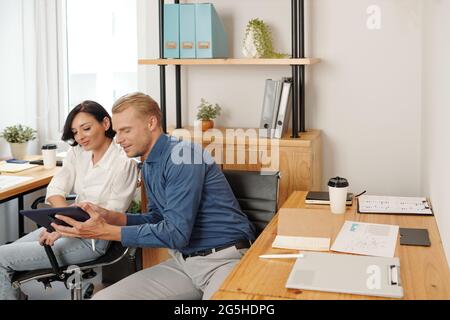 Smiling young business colleagues watching funny videos or memes on tablet computer instead of working on project Stock Photo