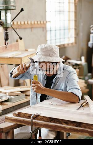 Serious young carpenter working in workshop and carving wood with chisel and hammer Stock Photo