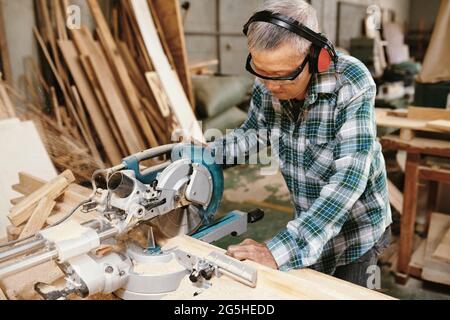 Experienced elderly carpenter in protective goggles and earmuffs cutting wooden planks with circular saw Stock Photo