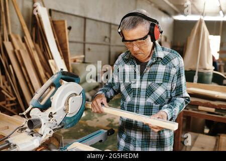 Serious senior carpenter measuring wooden plank before cutting it with circular saw Stock Photo
