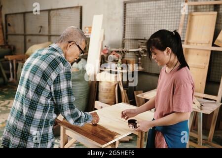 Smiling teenage girl looking at her grandfather covering surface of wooden board with special oil Stock Photo