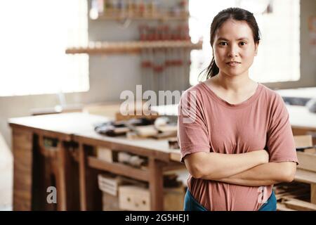 Portrait of young female carpenter crossing arms and looking at camera when standing in workshop Stock Photo