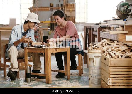 Joyful young carpenters enjoying coffee break in workshop, they are discussing picture of creative woodworking project on smartphones Stock Photo