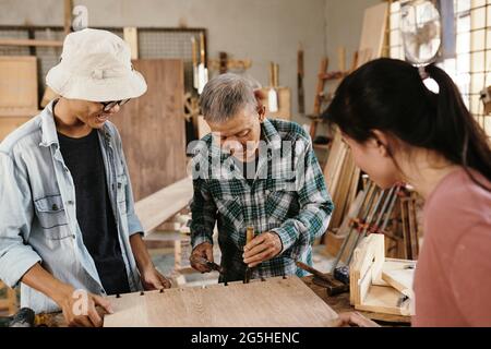 Young people watching experienced elderly carpenter using chisel and hammer when making cuts in wooden board to fit in butterfly inlay keys Stock Photo