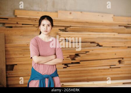 Portrait of young serious female carpenter standing in workshop against pile of wooden planks and looking at camera Stock Photo