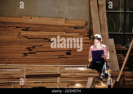 Concentrated young carpenter sitting on stack of wooden planks in sunrays and drawing or writing on clipboard Stock Photo